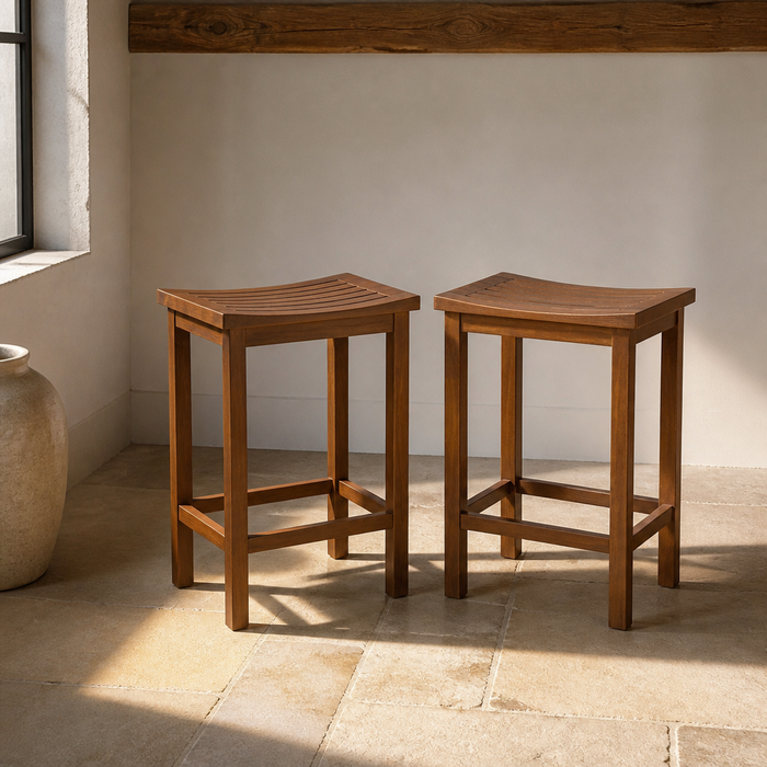 Set of two acacia wood counter height bar stools with saddle scooped slatted seats on a limestone flag floor