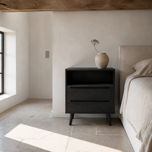 Black bedside nightstand beside a linen dressed bed on a limestone flag floor
