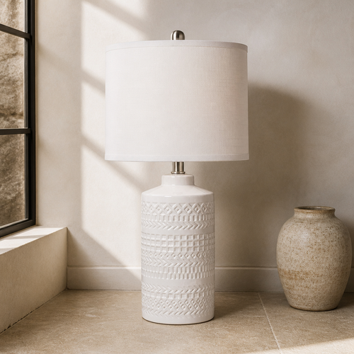 White ceramic table lamp with embossed geometric base and white linen drum shade on a limestone floor