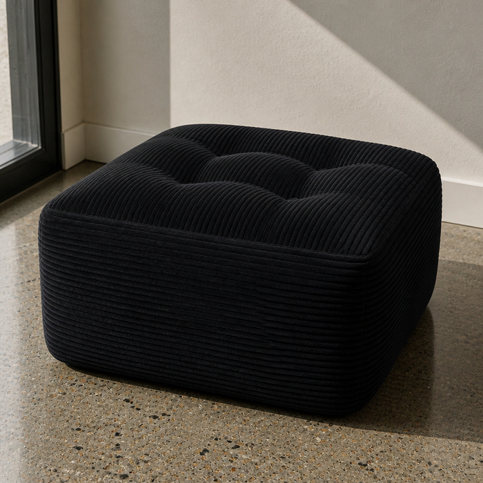 Square black corduroy floor pouf, close product portrait in a bone plaster room